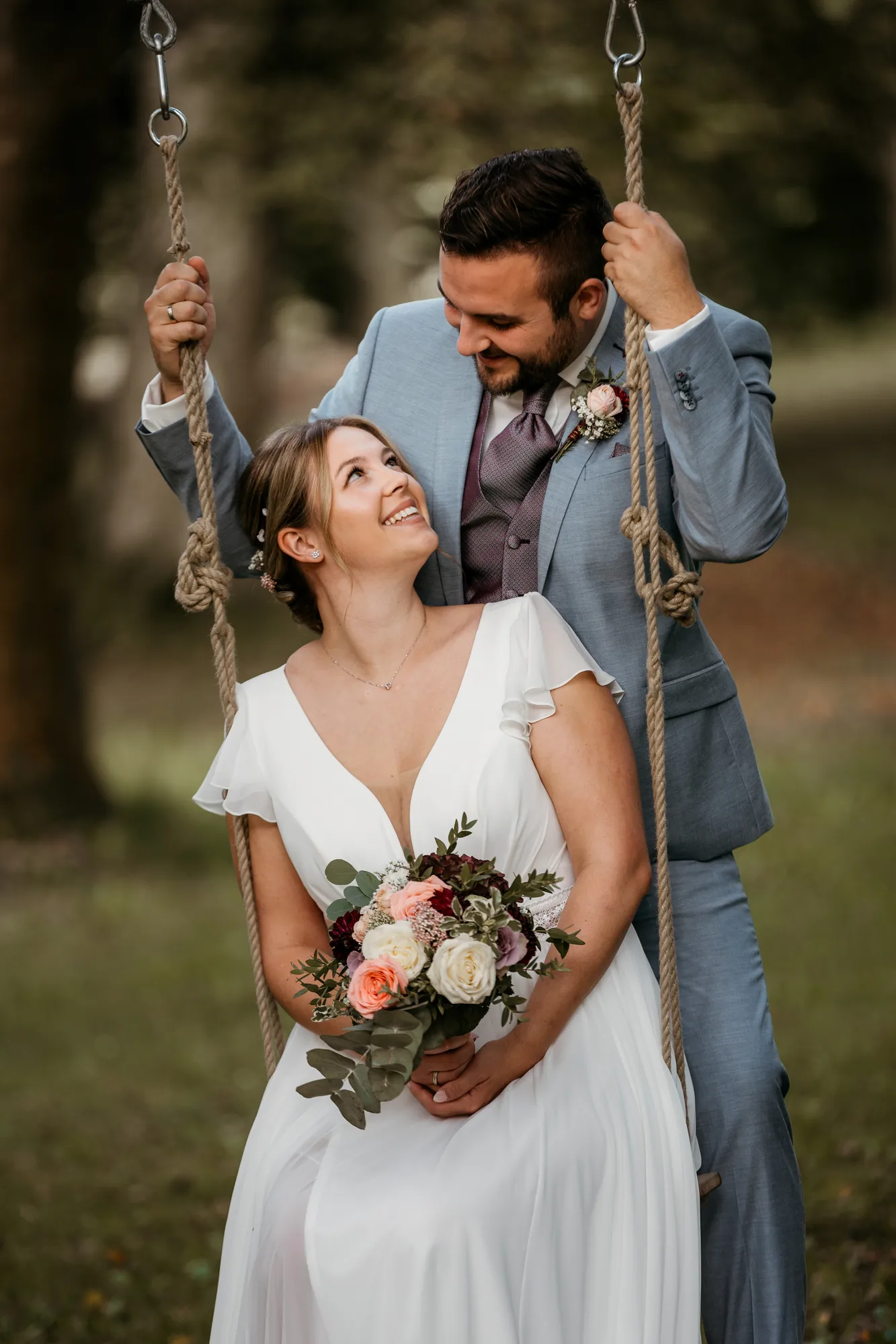 Braut auf einer Holzschaukel, der Bräutigam dahinter – Hochzeitsfotograf aus Niederbayern