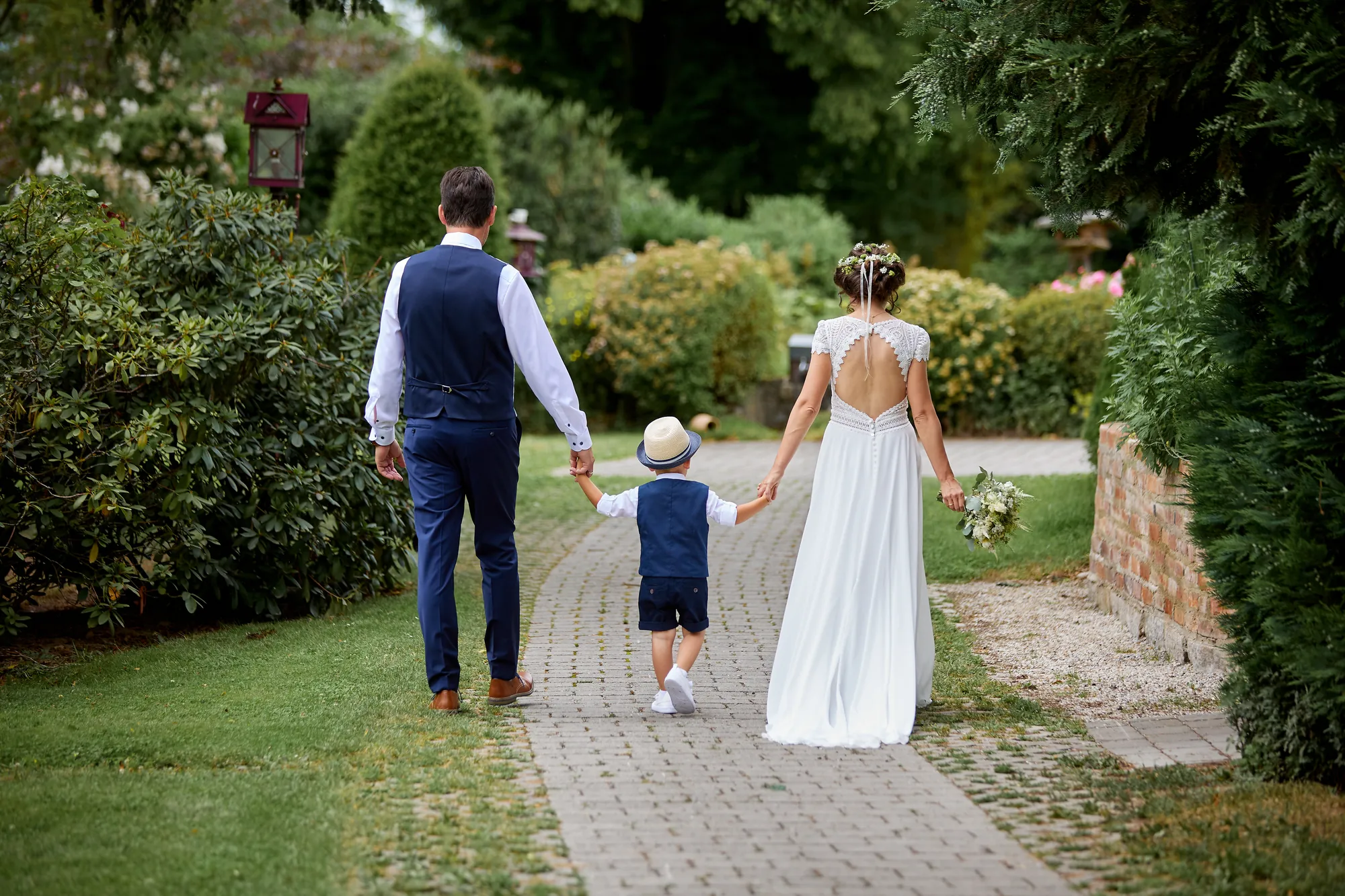 Brautpaar mit Kind auf dem Weg durch den Garten, von hinten – Hochzeitsfotos von Reinhard Fürstberger